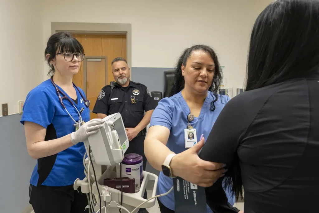 Nurse wrapping blood pressure reader around patient's arm, police officer in the background