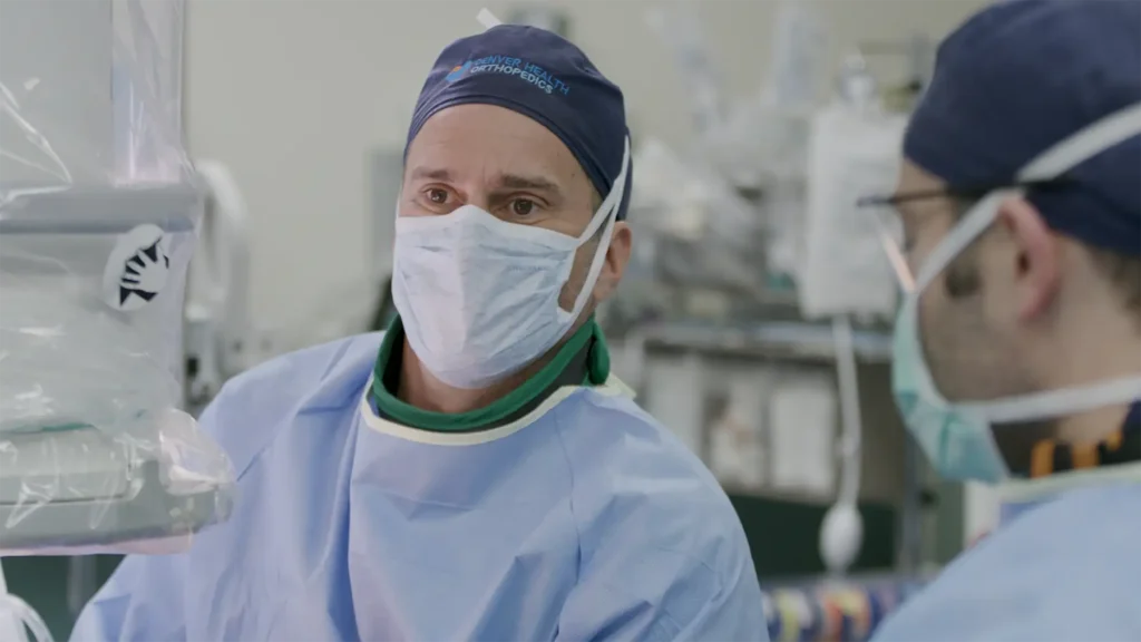 Doctor in surgery with a face mask, scrubs, and cap that says "Denver Health Orthopedics"